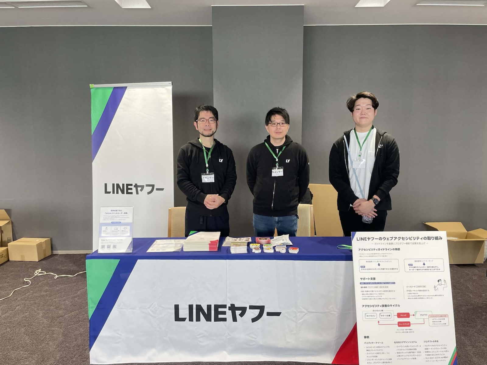 LINEヤフースポンサーブースで撮影された現地参加メンバーの写真。手前にはLINEヤフーと書かれた幕が敷かれた机があり、卓上にノベルティやペーパーが並んでいる。その前に掲示用のポスターが立てかけてある。ブースの後ろにはLINEヤフーの中野、富田、慶島の三人がならんで立っている