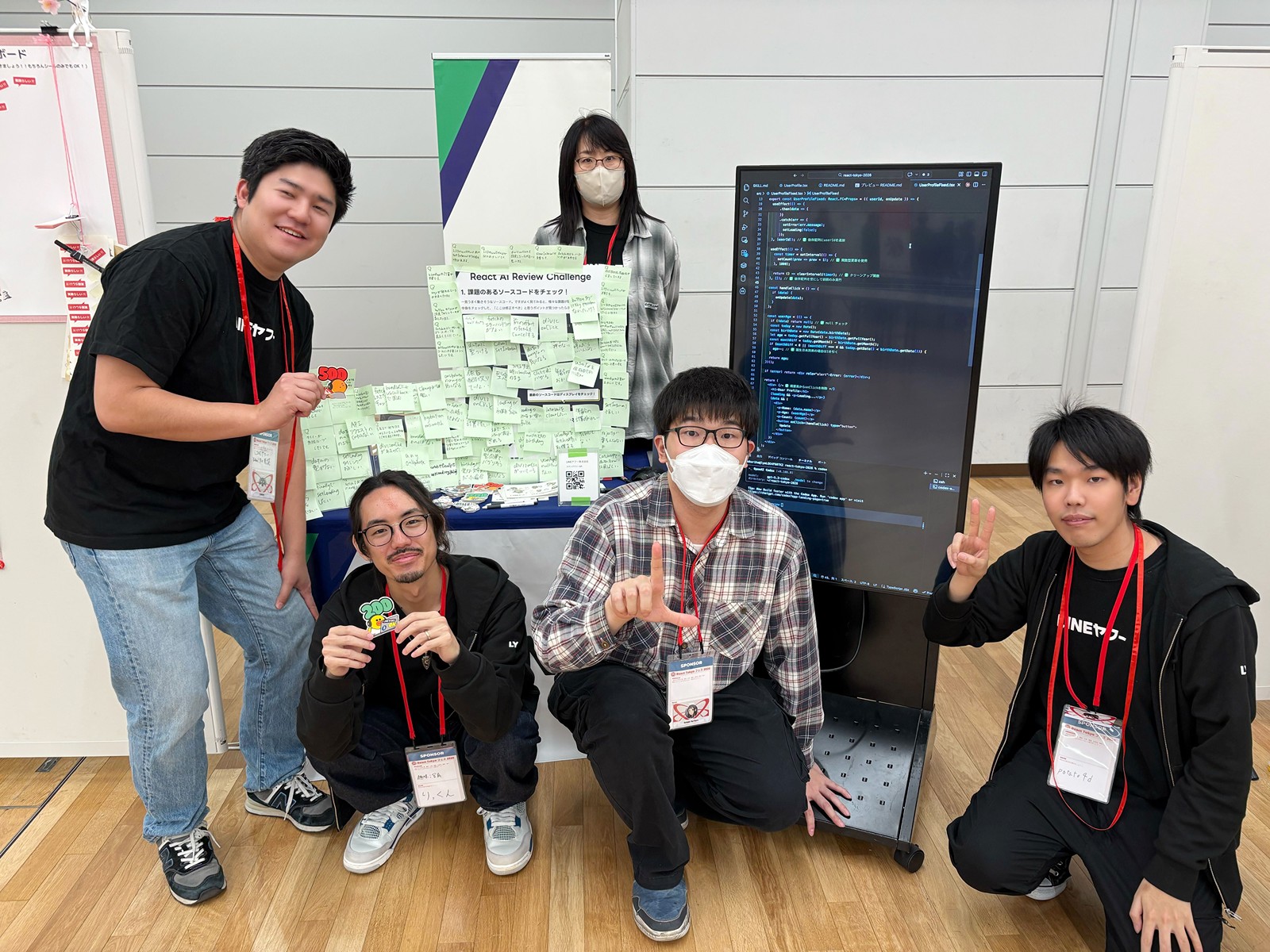 l1lhu1hu1、sezawa、りっくん、kokuriha、potato4dの集合写真である。それぞれLINEヤフーのロゴがあしらった服装やノベルティを所持して自社をアピールしている。背後には「React AI Review Challenge」と書かれたボードがあり、緑色の付箋が多数貼られている。右側にはコードが表示された大型モニターが設置され、会場内の木目調の床とパネル壁が見える展示ブースの様子である。
