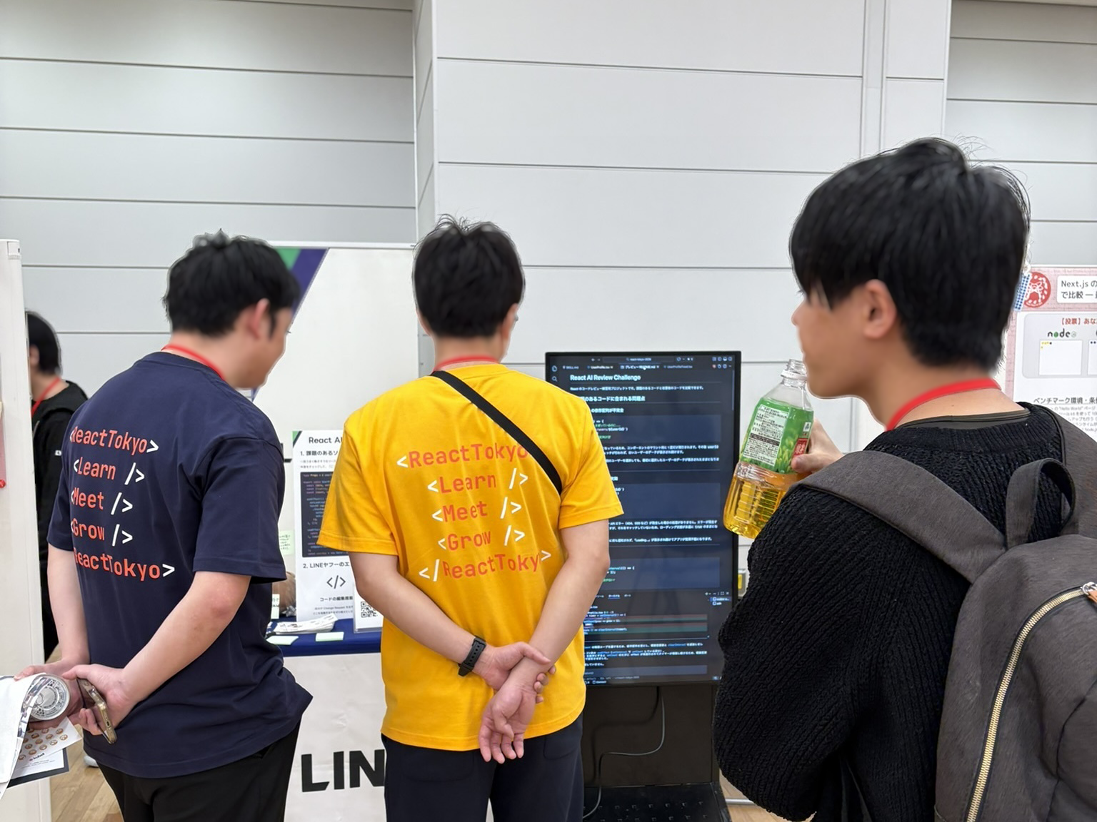 3人の来場者が展示ブースの大型縦型モニターを見ている後ろ姿の写真である。中央の人物は黄色のReactTokyoのTシャツを着ている。両手を後ろで組み、モニターに表示されたコードや説明文を見ている。左の人物は紺色のReactTokyoのTシャツを着ている。右の人物はペットボトル飲料を手に持ちながら画面を見ている。モニターには「React AI Review Challenge」の画面とソースコードが表示されている。手前には「LINEヤフー」と書かれたテーブルクロスが見え、屋内会場のパネル壁と木目調の床が背景に広がっている展示風景である。