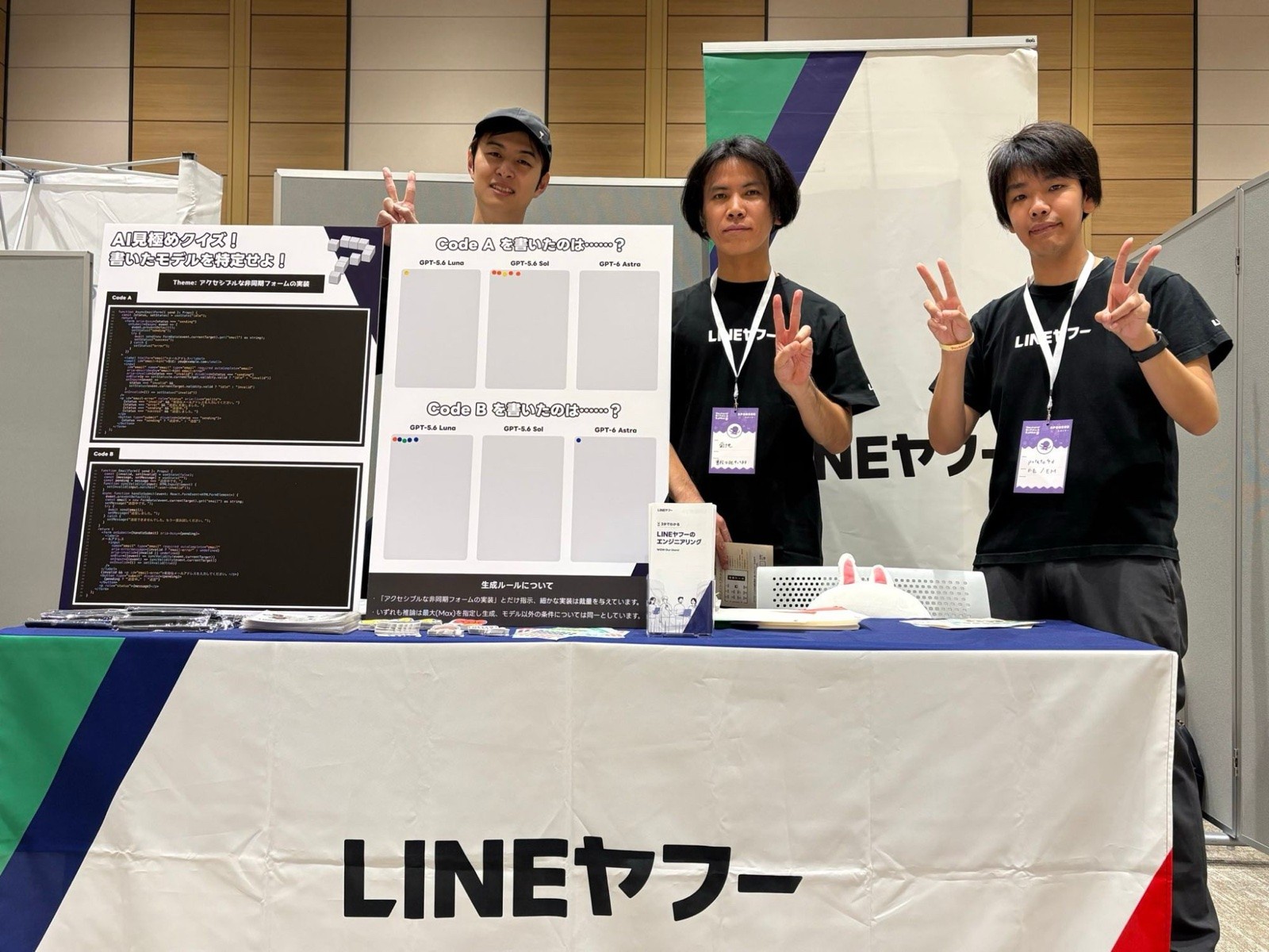 LINEヤフーの展示ブースで撮影した3人の集合写真である。左の人物はキャップをかぶり、パネルの後ろからピースサインをしている。中央と右の人物は「LINEヤフー」のロゴ入りの黒いTシャツを着て名札を下げ、ピースサインをしている。手前には「LINEヤフー」と大きく印字された白いテーブルクロスをかけた机があり、左側に「AI見極めクイズ！書いたモデルを特定せよ！」のパネルと、Code AとCode Bを書いたモデルを投票するパネルが立てられている。机の上には冊子やステッカー類が並び、背後には緑と紺の斜めラインが入ったLINEヤフーのバナーが立っている。