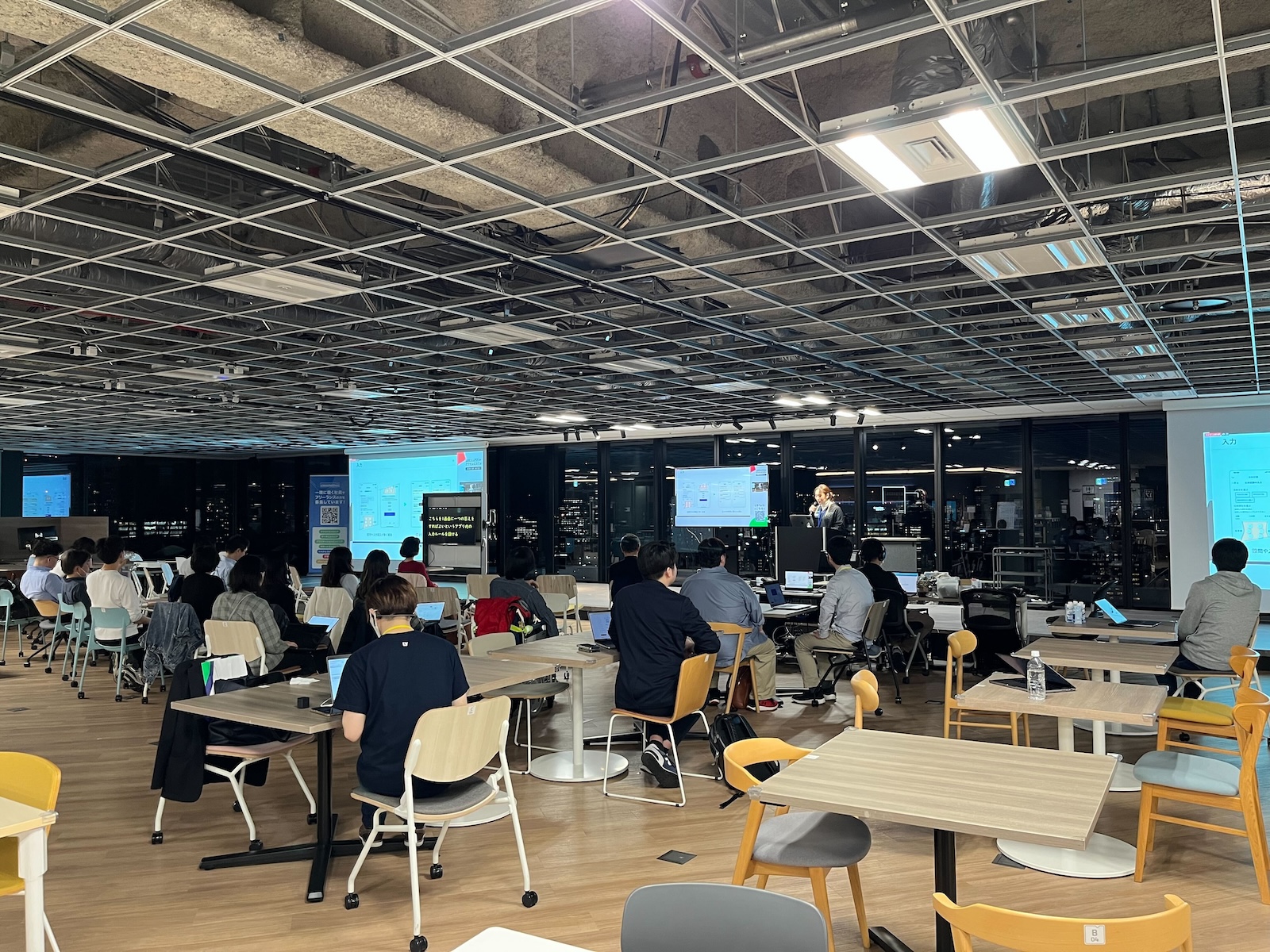勉強会の会場の風景。フロアに椅子が30脚ほど並べられていて、来場者が座っている。壁一面の大きな窓を背に、登壇者が立っていて大きなディスプレーにスライドを表示して話をしている。登壇者の左右にはスクリーンがあってスライドが映されている。天井はパネルがなく開放感のあるスペースになっている。